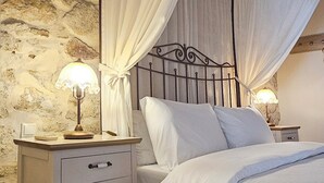 Un dormitorio con pared de piedra, cama con ropa de cama blanca, mesita de noche con lámpara y dosel.