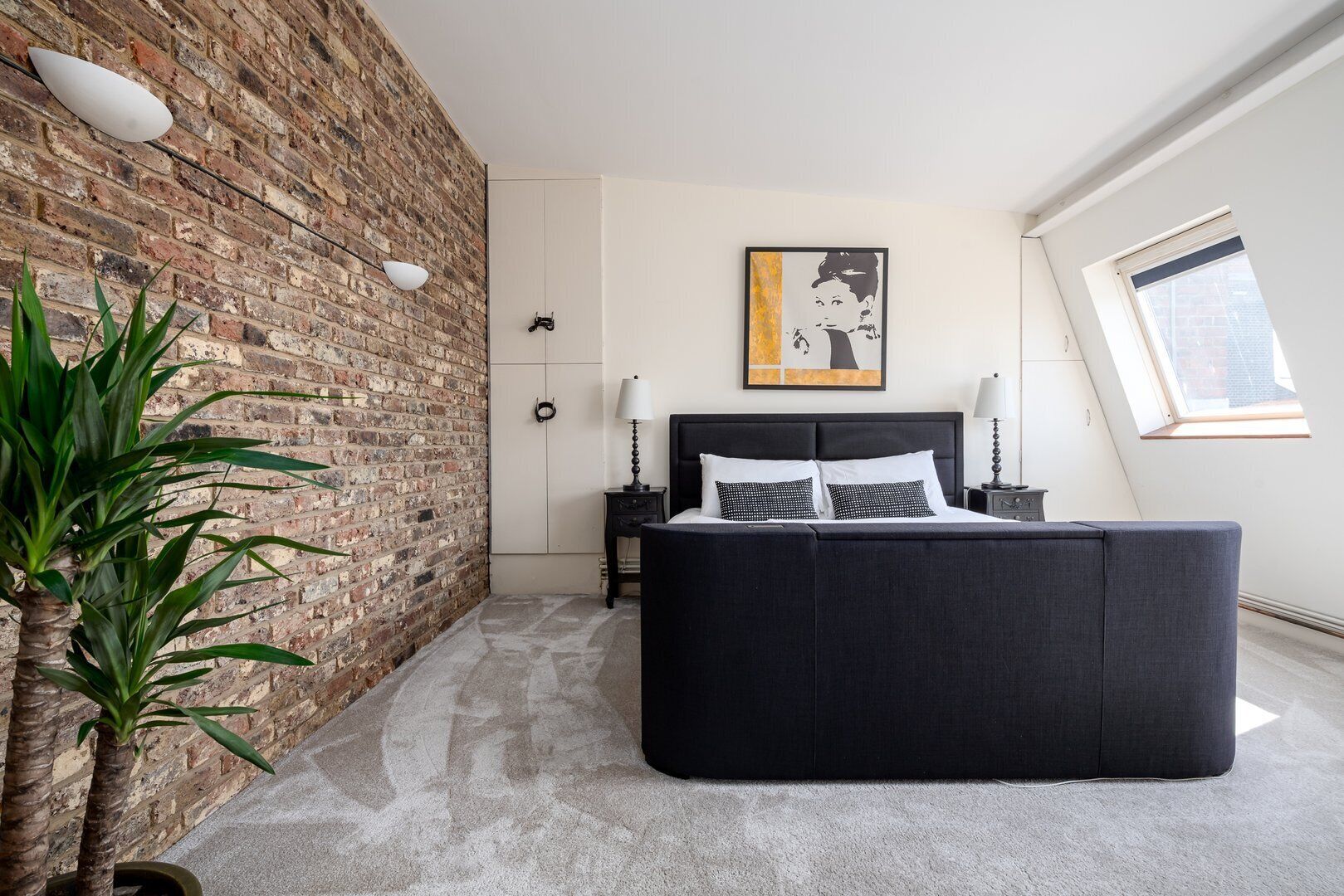 A bedroom with a brick wall, a bed with black bedding, a nightstand, a lamp, and a window with a view.