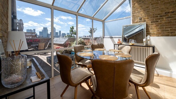 A modern dining area with a glass table and chairs, a city view, and a brick wall. - GuestReady - Modern 2BR w Balcony in Poplar (London)