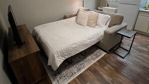 A bedroom with a bed, bedside table, lamp, and a view of a kitchen area. - Downtown Starkville Apartment! (Starkville)