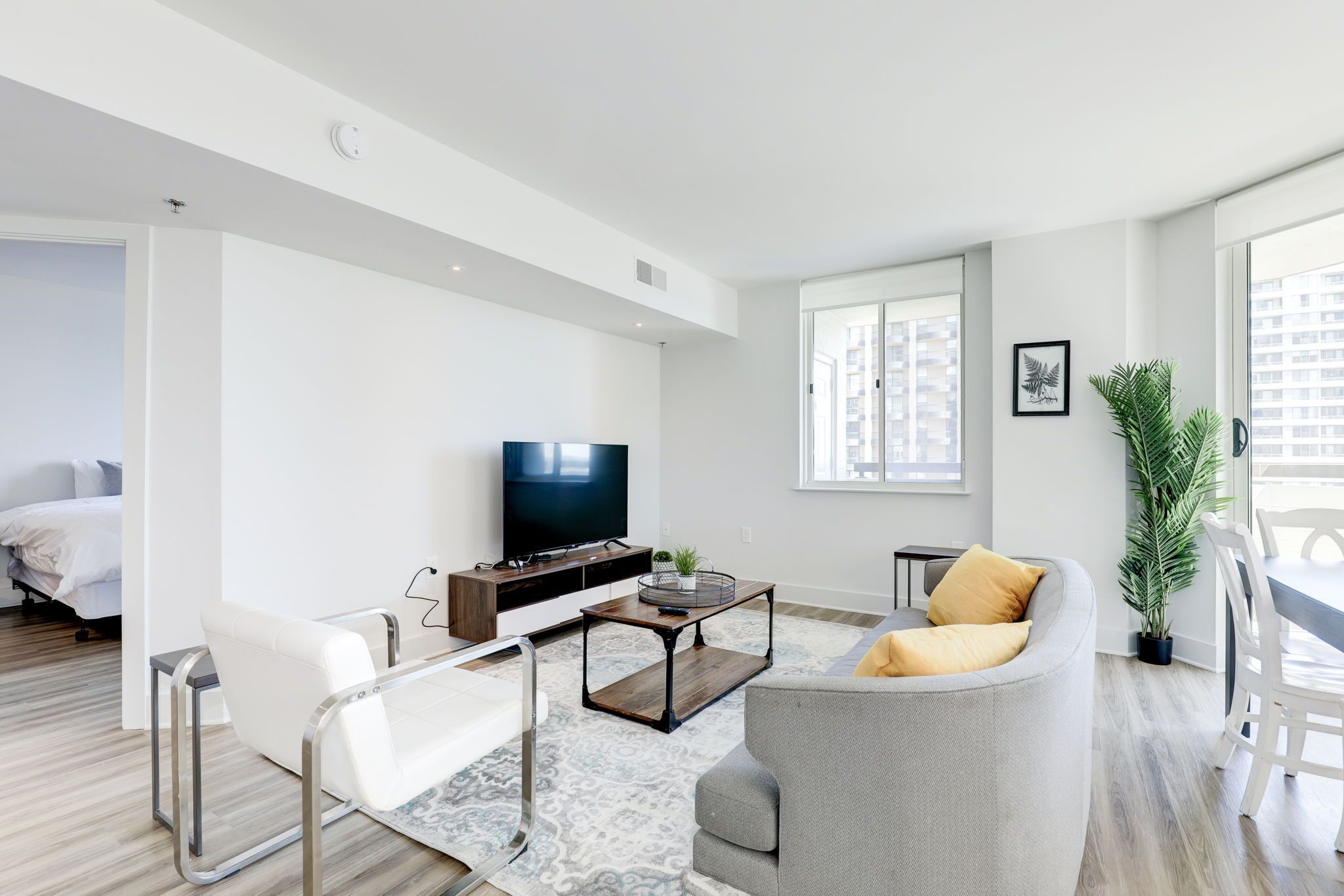 A modern living room with a sofa, a coffee table, and a TV. There is a window with a view of buildings, a potted plant, and a bed in the background.
