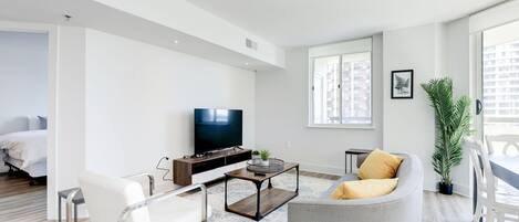 A modern living room with a sofa, a coffee table, and a TV. There is a window with a view of buildings, a potted plant, and a bed in the background.