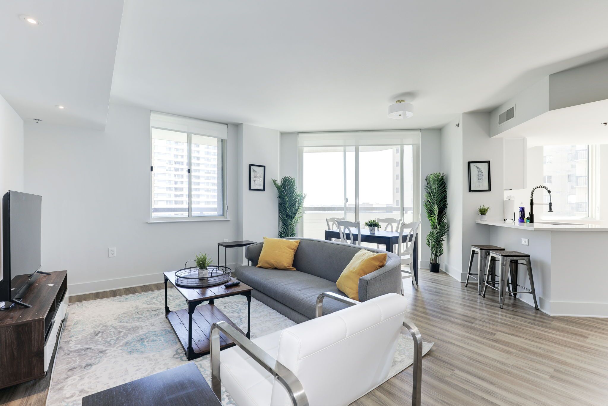 A modern living room with a grey sofa, white armchair, and a coffee table. There is a dining area with a table and chairs, and a kitchen area with a sink and stools.