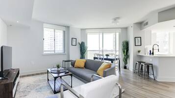 A modern living room with a grey sofa, white armchair, and a coffee table. There is a dining area with a table and chairs, and a kitchen area with a sink and stools.