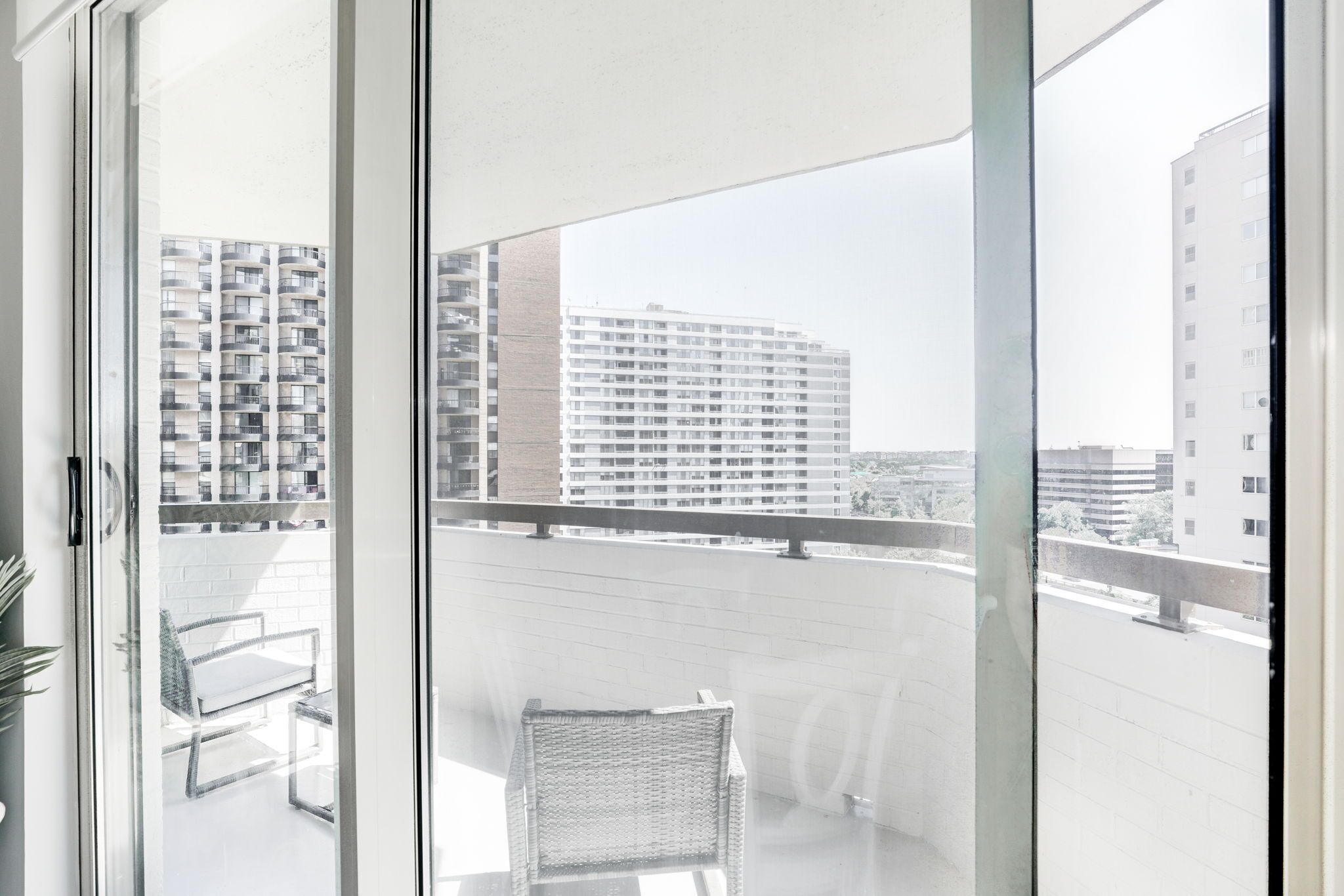 A balcony with a wicker chair and a table, overlooking a cityscape with high-rise buildings.