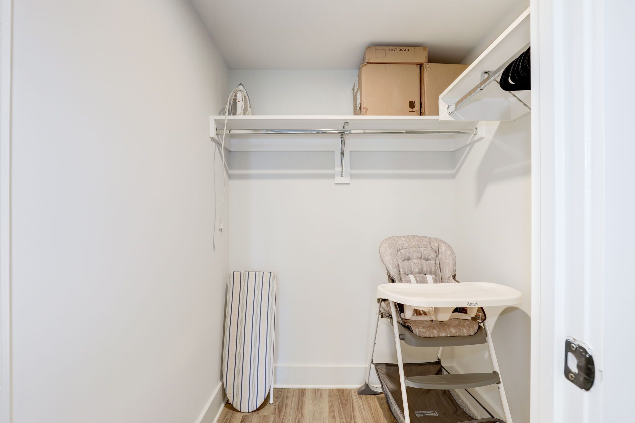 A compact room with a white wall, a small ironing board, a baby high chair, and a shelf with boxes.