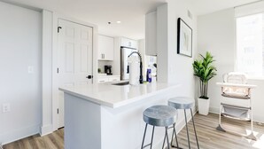 Une cuisine moderne avec des meubles blancs, un îlot central doté de tabourets et une vue sur le salon, agrémenté d’une plante et d’une fenêtre.