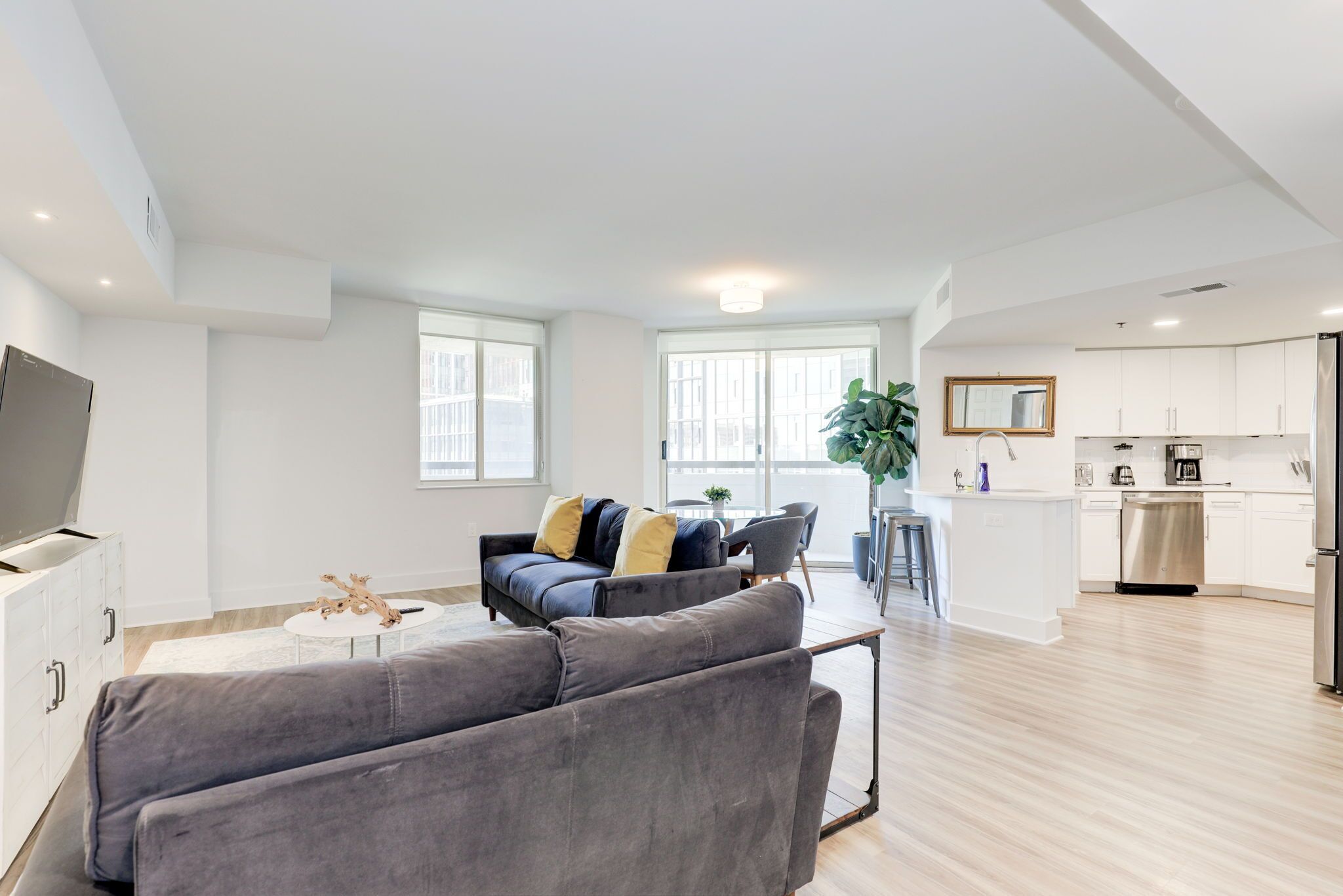 A modern living room with a grey sofa, a coffee table, and a TV. There is a kitchen area with white cabinets and a stainless steel refrigerator.