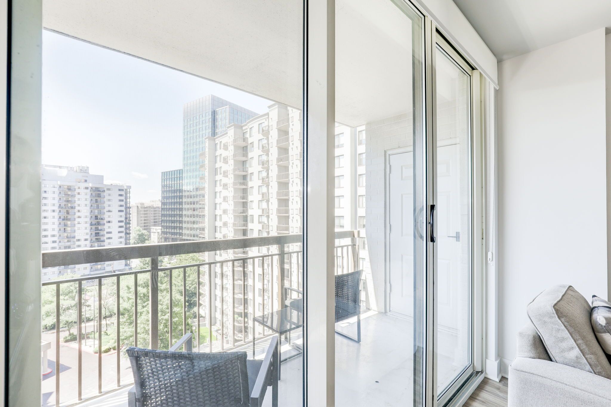 Een balkon met een glazen deur, een bank en een stoel, met uitzicht op een stadslandschap met hoge gebouwen.