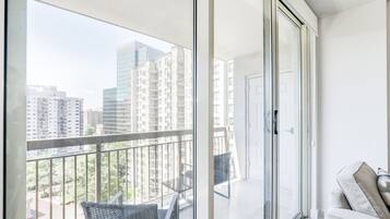 Een balkon met een glazen deur, een bank en een stoel, met uitzicht op een stadslandschap met hoge gebouwen.