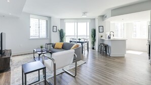 Un salon moderne avec un canapé, une table basse et un téléviseur. Il y a également un coin repas avec une table et des chaises. La cuisine est équipée d’un comptoir et de tabourets.