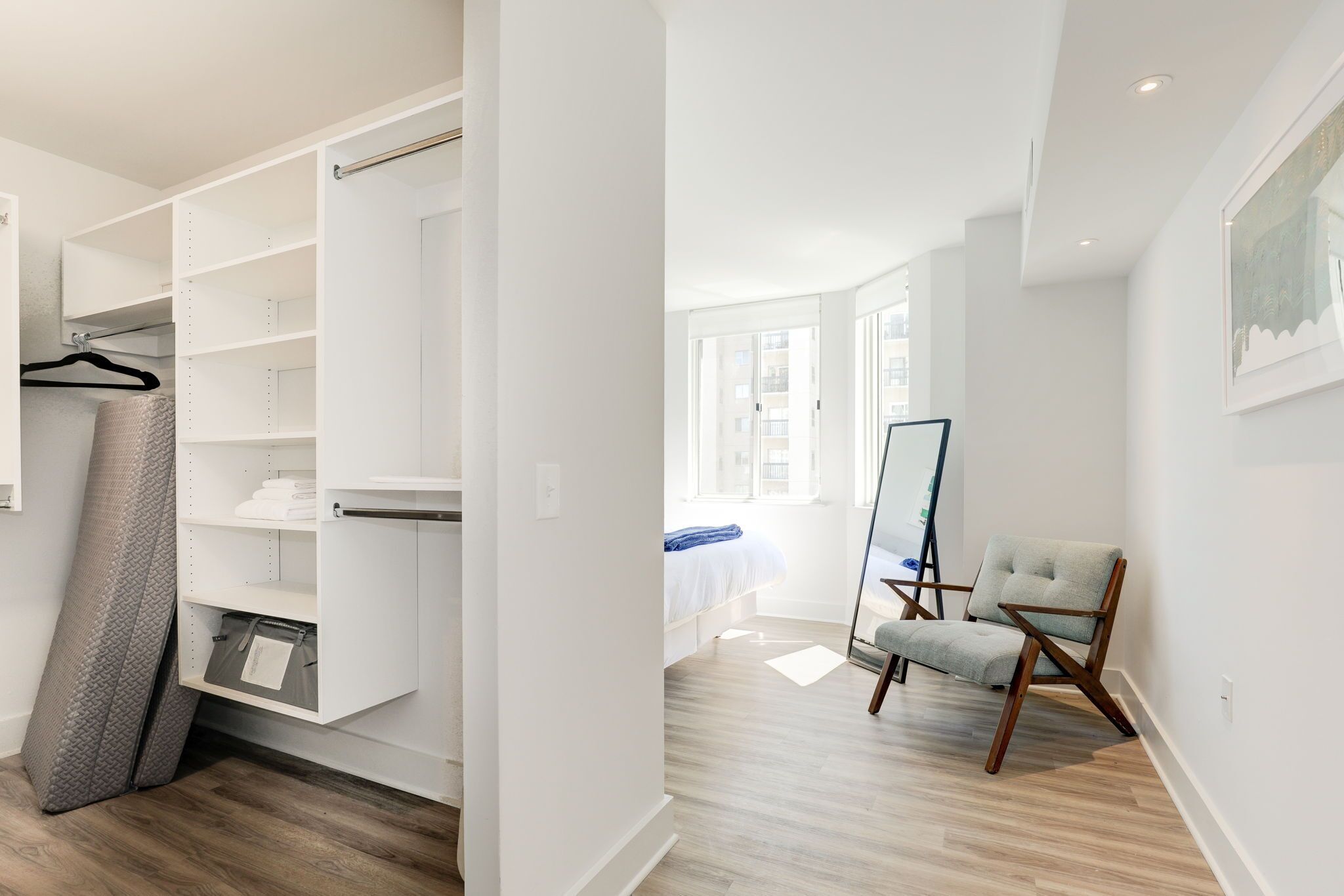 Een moderne slaapkamer met inloopkast, een bed met een blauwe deken, een stoel en een groot raam.