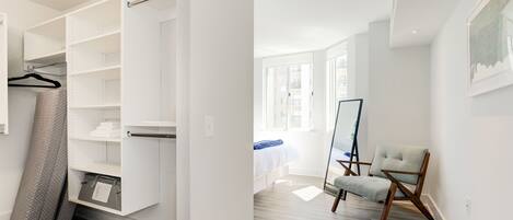 A modern bedroom with a walk-in closet, a bed with a blue blanket, a chair, and a large window.