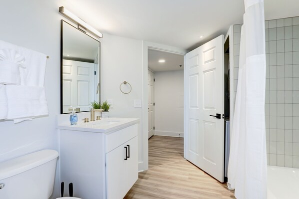 Un baño moderno con lavamanos, espejo y retrete blancos. Hay una cortina de ducha y una planta en el mueble de la lavamanos.