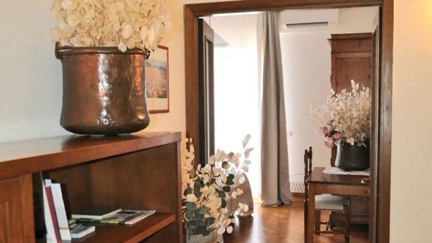 Ein Bücherregal aus Holz mit Büchern, ein Metalltopf mit Blumen und ein Teppich auf einem Holzboden.