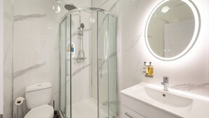 Uma casa de banho moderna com cabine de duche envidraçada, lavatório branco com espelho e paredes em mármore.