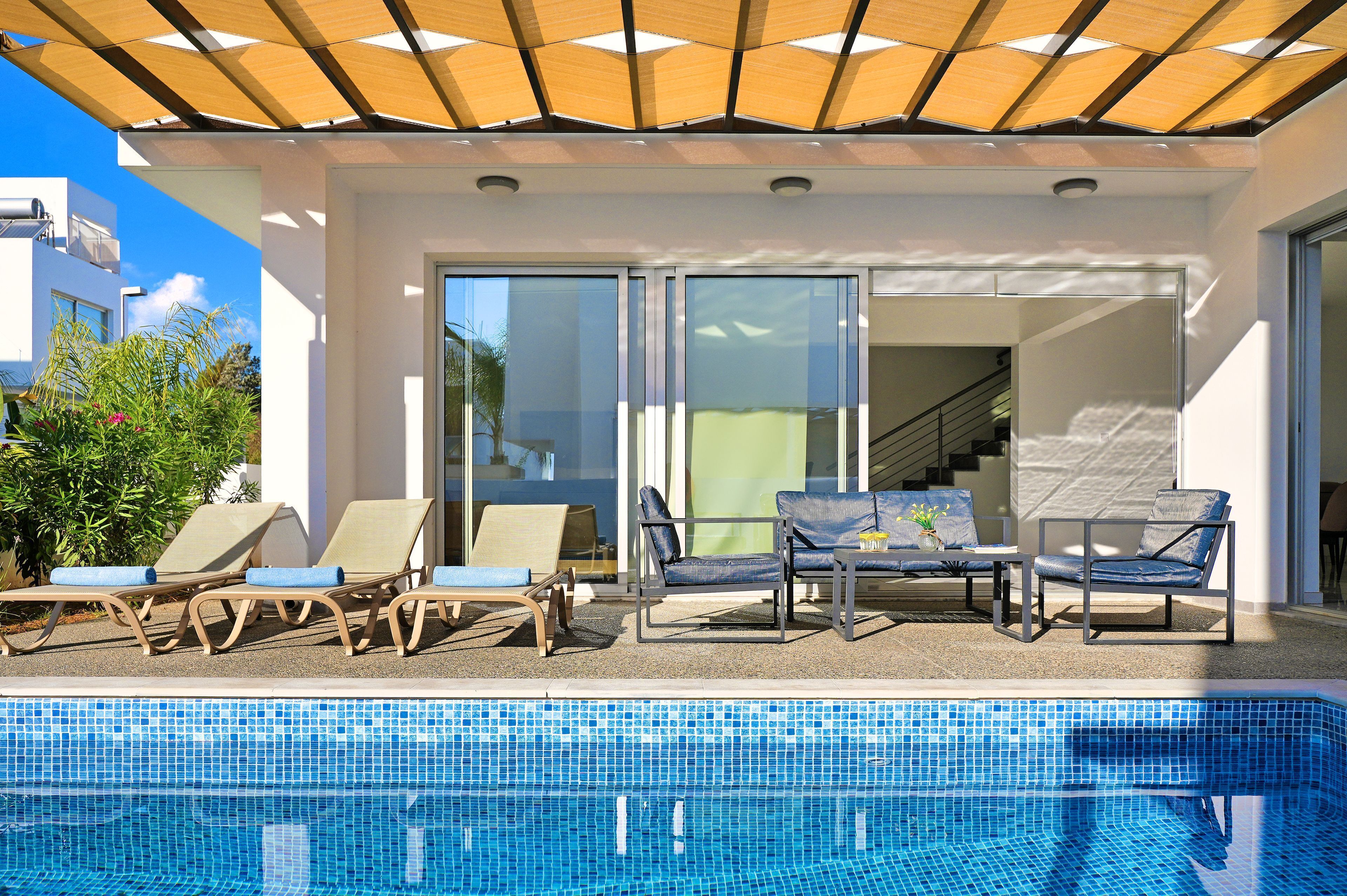 Un espace aménagé au bord de la piscine, avec des chaises longues et une table, un coin salon couvert et une vue sur un escalier menant à un niveau supérieur.