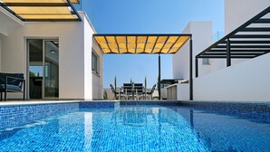 Una piscina moderna al aire libre con superficie azulejada azul, una zona de estar cubierta con mesa y sillas, y un cielo despejado al fondo.