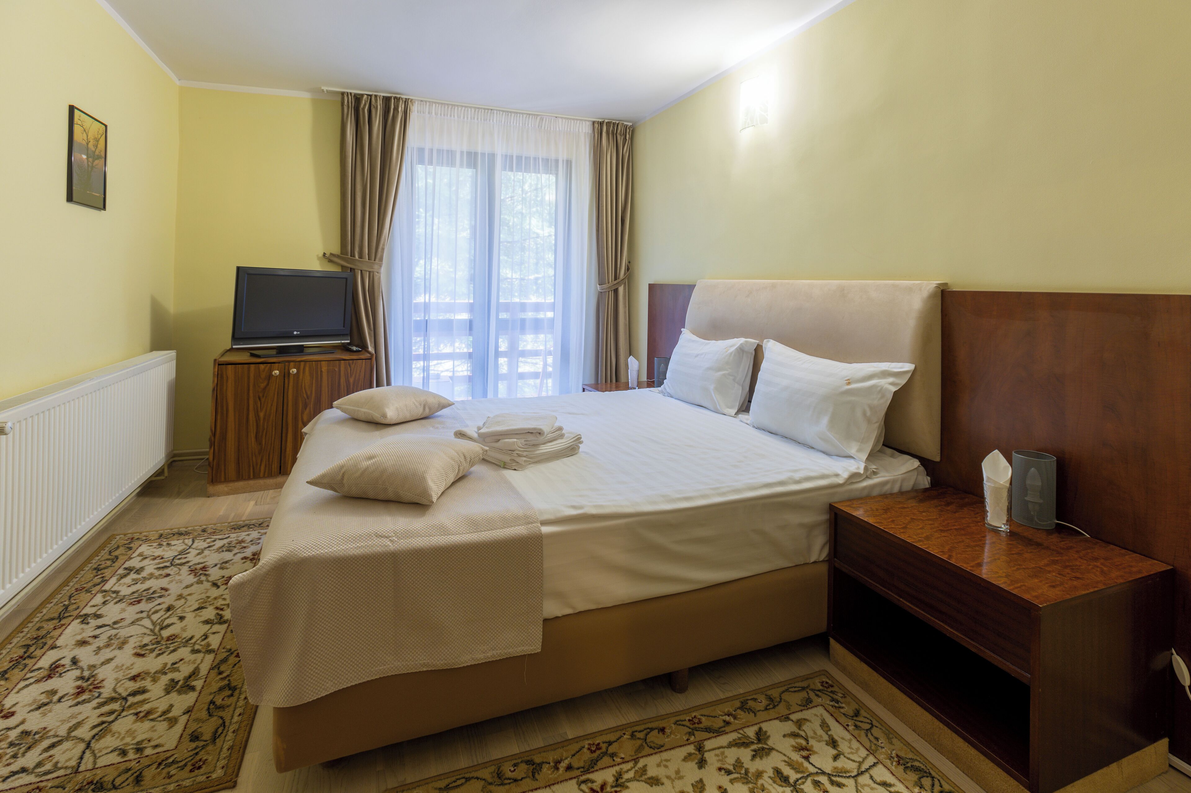 Une chambre d’hôtel avec un lit, un téléviseur posé sur une commode en bois, une table de chevet et une fenêtre avec des rideaux.