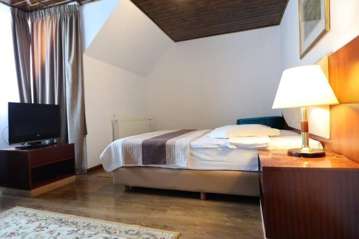 Un dormitorio con cama, mesita de noche con lámpara, televisor sobre un mueble bajo y ventana con cortinas.