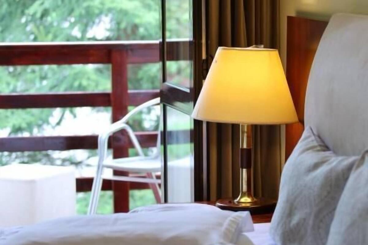 Habitación de hotel con cama, almohadas, lámpara de noche y vista a zonas verdes por la ventana.