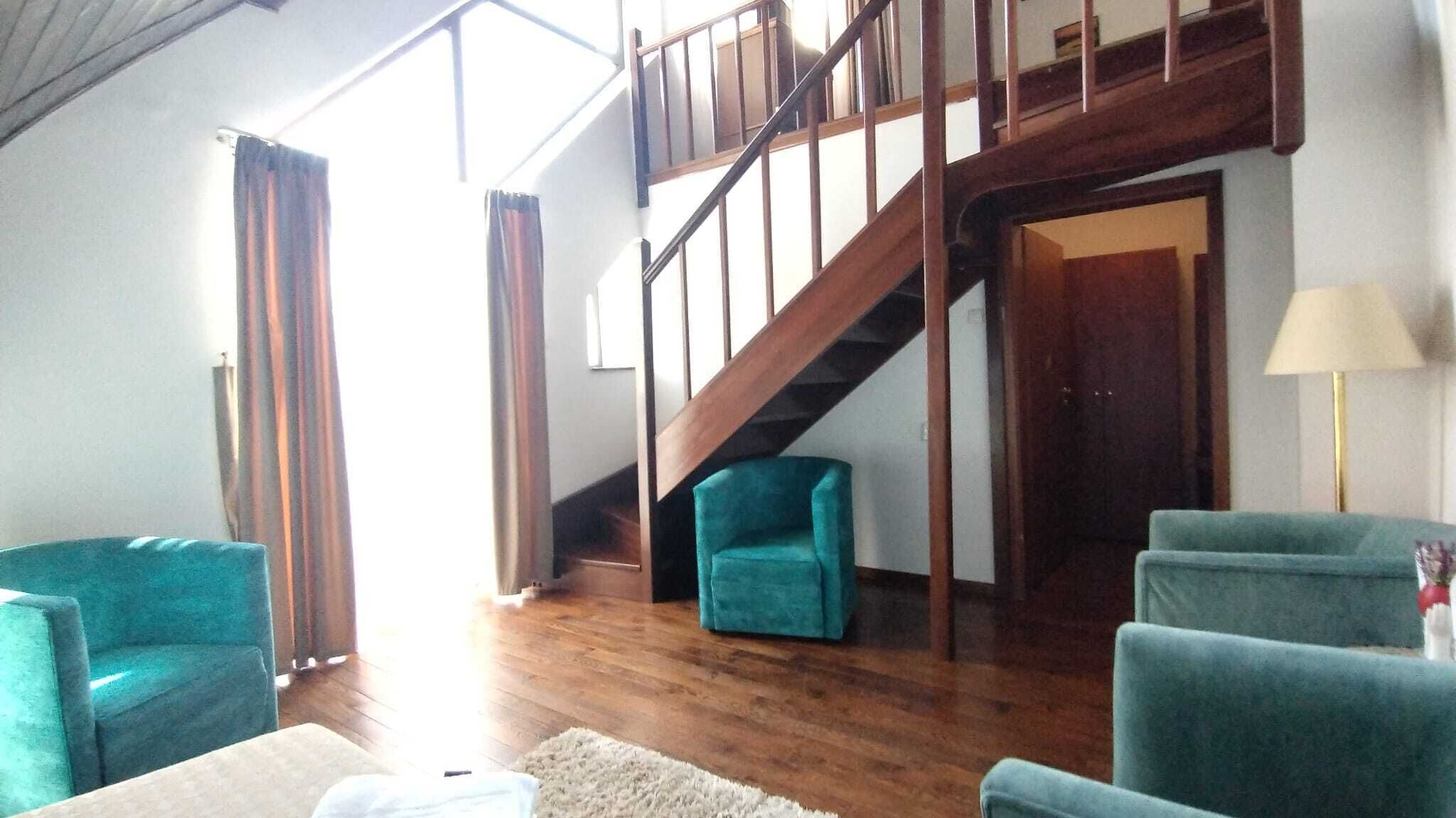 A spacious living area with wooden flooring, a staircase, and teal armchairs.