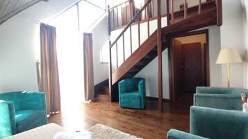 A spacious living area with wooden flooring, a staircase, and teal armchairs.