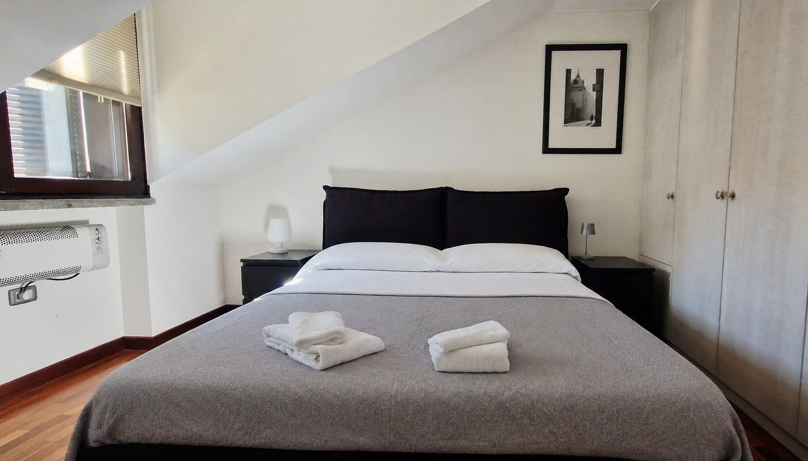 Una cama bien hecha con ropa de cama blanca, un edredón gris y dos toallas dobladas sobre la cama. Se observa un cabecero negro, una mesita de noche con una lámpara y un cuadro en la pared.