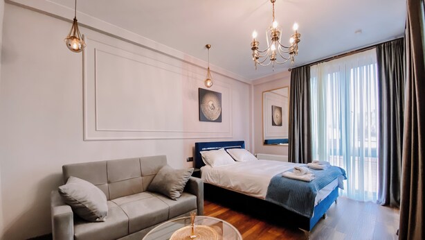 Una habitación de hotel moderna con un sofá gris, una cama con cabecera azul, una mesa de centro redonda de cristal y una araña.