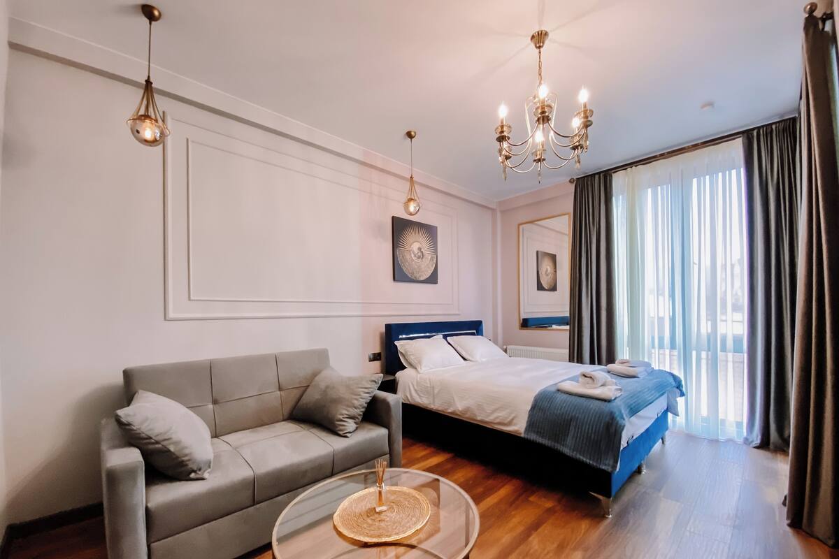 A modern hotel room with a grey sofa, a bed with a blue headboard, a round glass coffee table, and a chandelier.