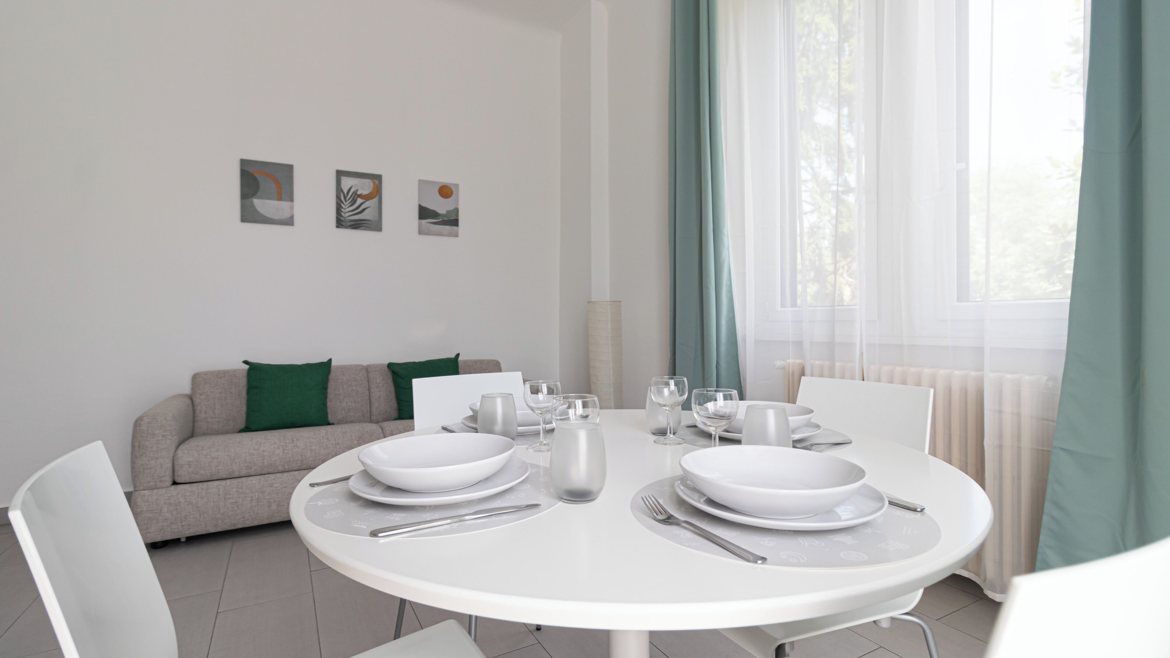 A modern dining area with a round table set for four, a grey sofa with green cushions, and three framed artworks on the wall.