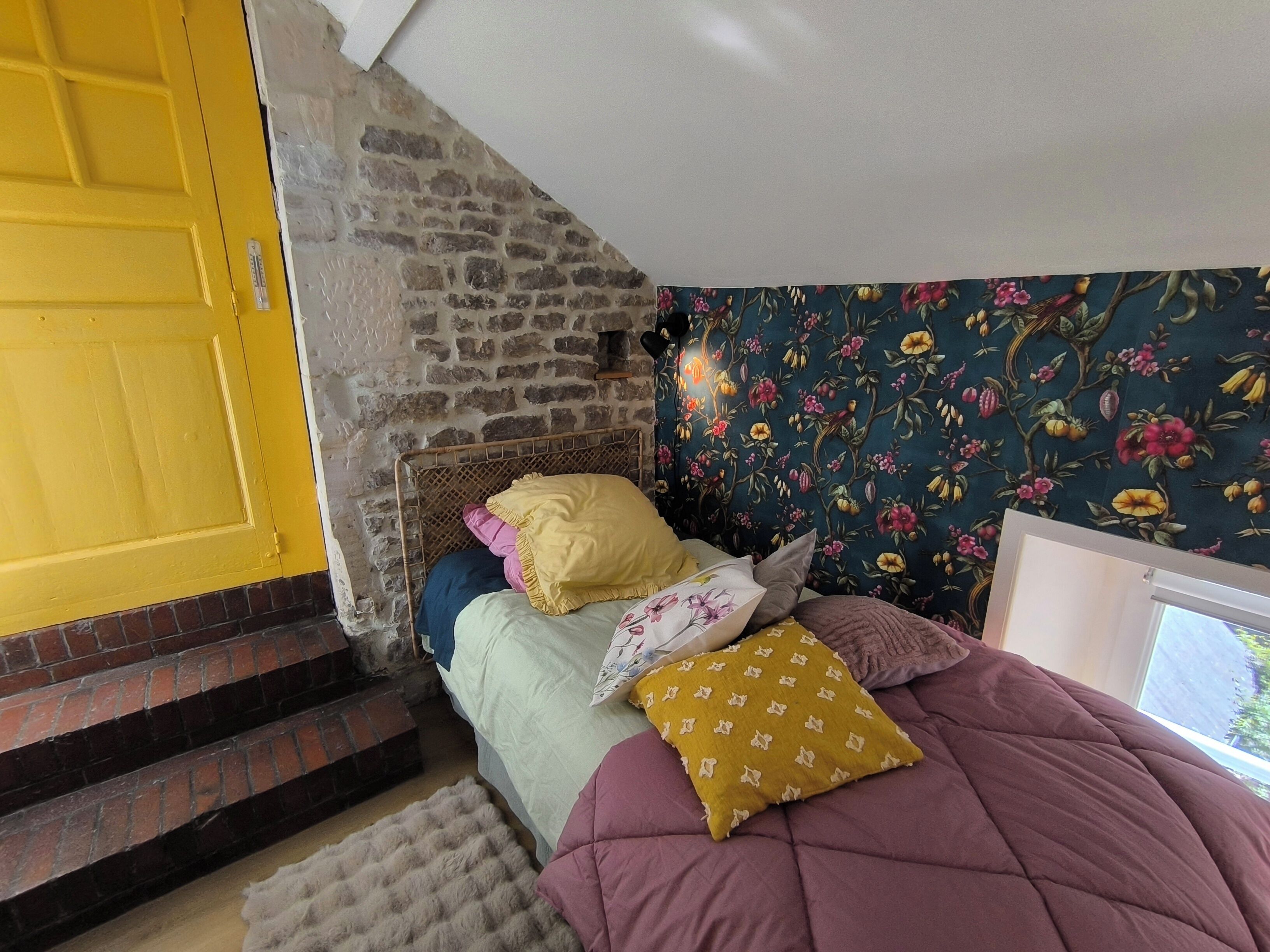 A bedroom with a stone wall, a yellow door, floral wallpaper, and a bed with multiple pillows.