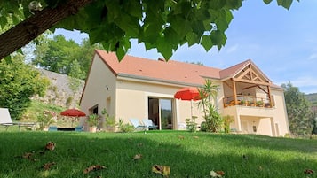 A two-story house with a red-tiled roof, a balcony with potted plants, and a grassy lawn with a few fallen leaves.