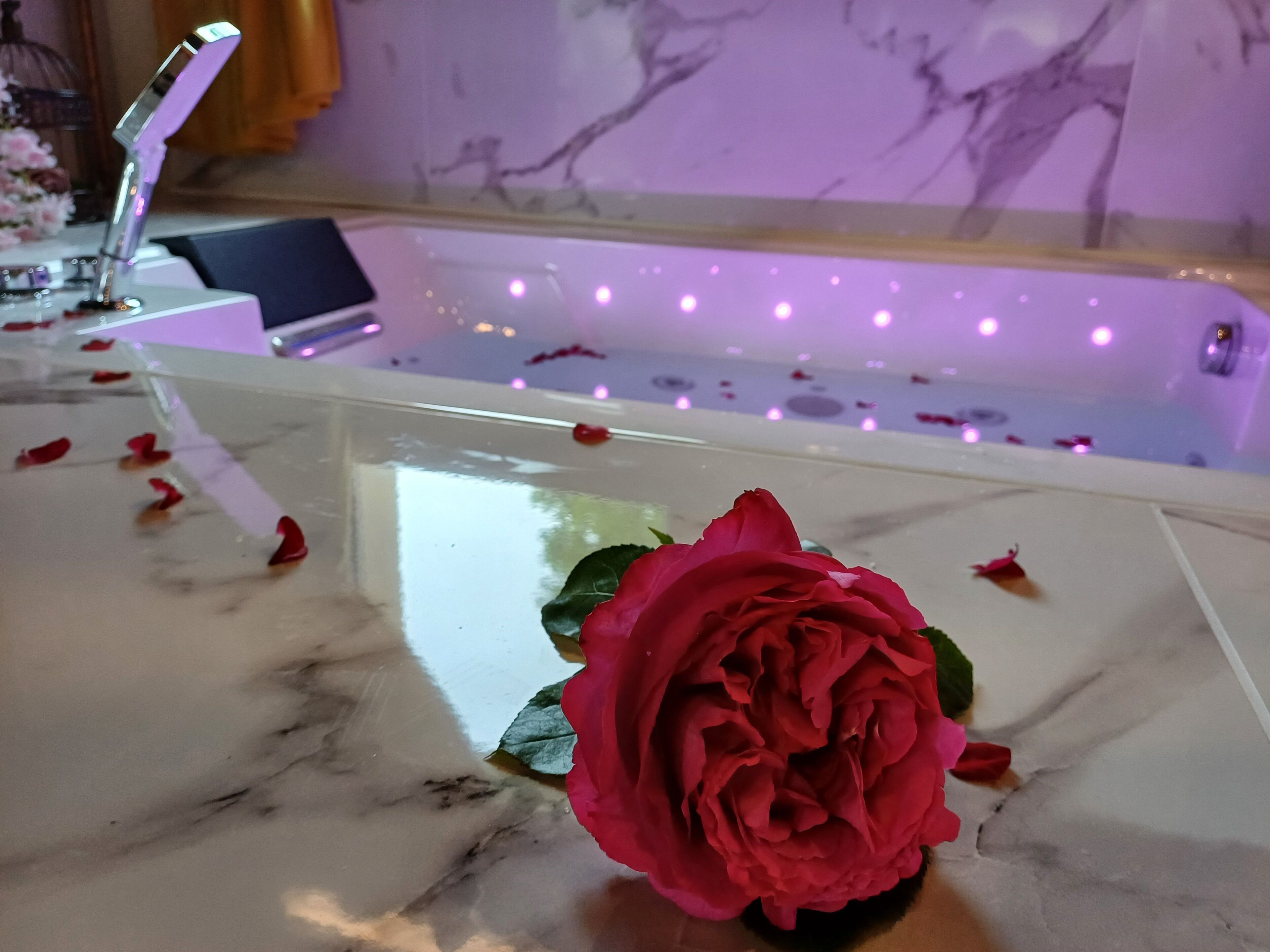 A marble-topped bathtub with rose petals scattered around, a rose in a vase, and a modern light fixture in the background.