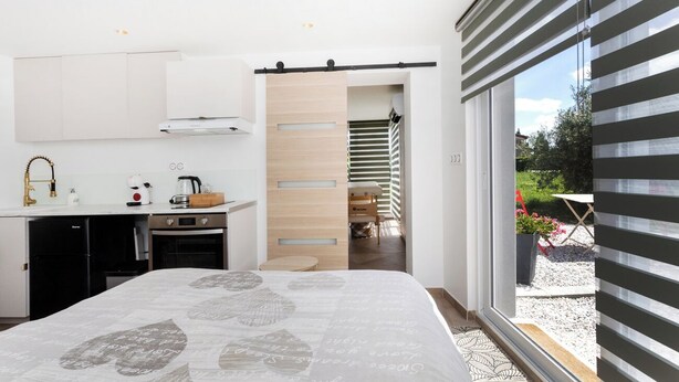 A modern bedroom with a large bed, a kitchen area with a dishwasher, and a sliding door leading to a balcony. - Escapade à 2 Bien Être et Déconnexion ! Jacuzzi Privatif (Roumens)