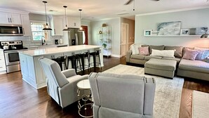 A spacious living room with a large gray sectional sofa, a white armchair, and a coffee table. A kitchen with white cabinets and a microwave is visible in the background. - Charming 3-bedroom secluded house in Acworth (Acworth)