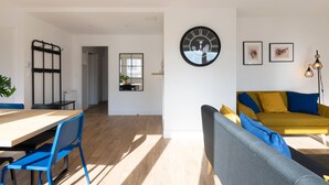 Ein modernes Wohnzimmer mit einer Couch, einem Esstisch und Stühlen. Eine große Wanduhr und gerahmte Kunstwerke sind ebenfalls vorhanden.