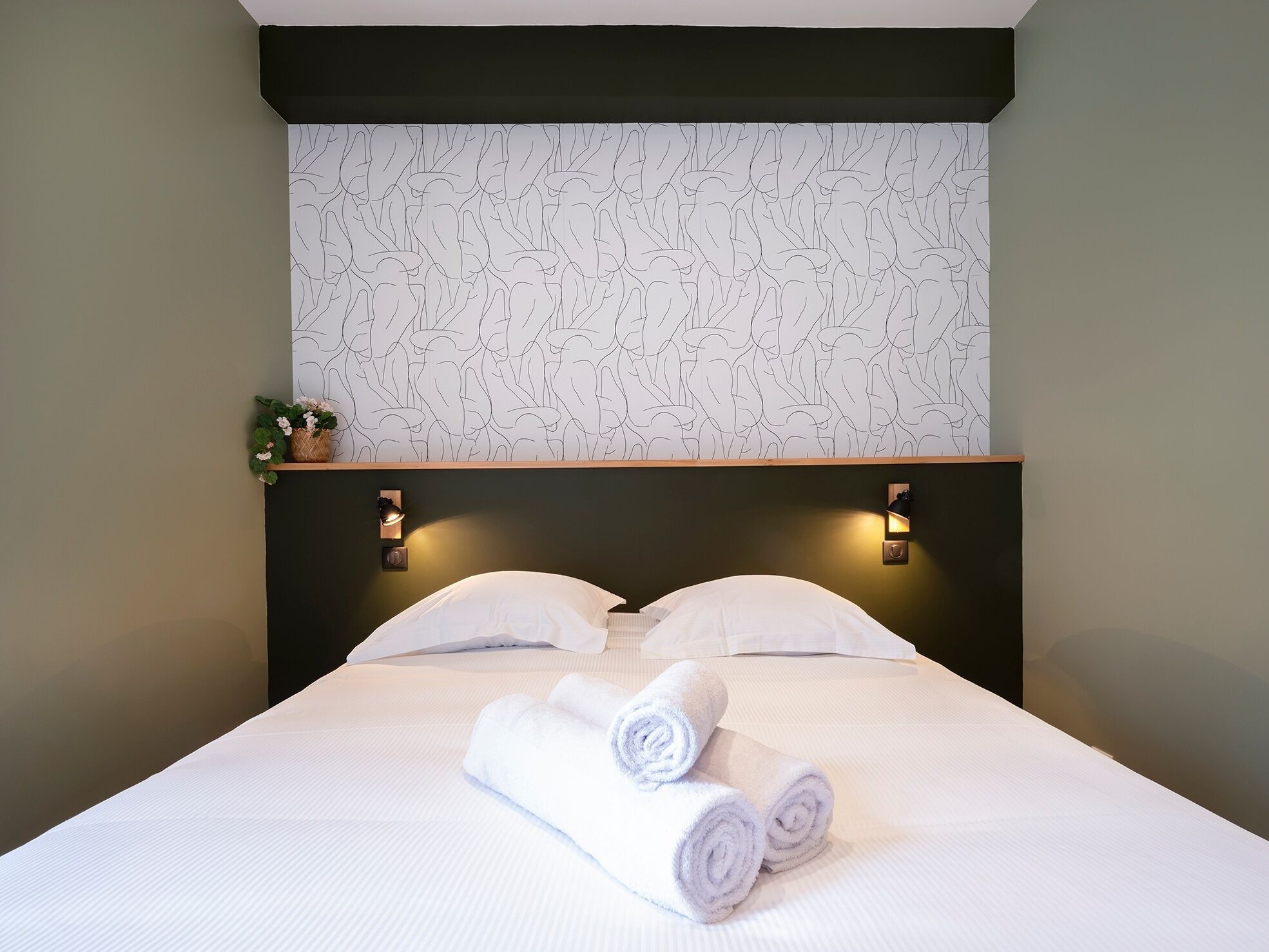 Un lit bien fait, avec deux oreillers et deux serviettes enroulées, un mur orné d’un motif de feuilles et deux appliques murales.