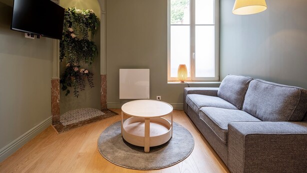 A modern living room with a grey sofa, a round wooden table, a flat-screen TV mounted on the wall, a window with a view of greenery, and a hanging lamp. - La Cachette de Stanislas - Centre-ville de Nancy (Nancy)
