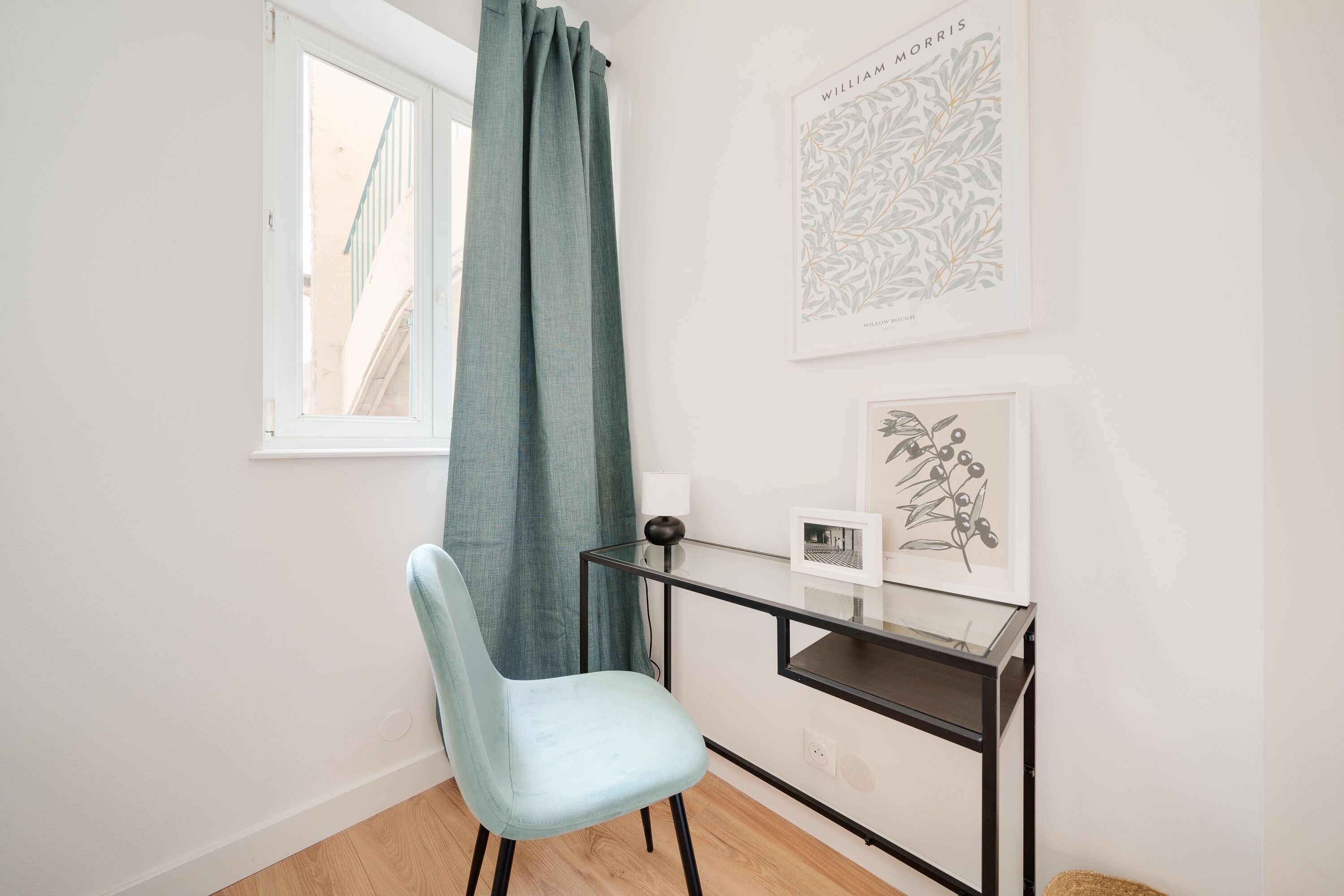A room with a light blue chair, a desk with a lamp, and two framed botanical prints on the wall.