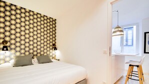 A bedroom with a patterned wallpaper, a bed with white bedding, two bedside lamps, and a view of a kitchen area with a stool and a hanging light fixture. - Le Cachet de la Craffe - Au cœur de Nancy (Nancy)