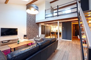 A modern living room with a large TV, leather sofa, and a dining area with a high ceiling and exposed beams. - Relais Ducal - Côté Duplex (Nancy)