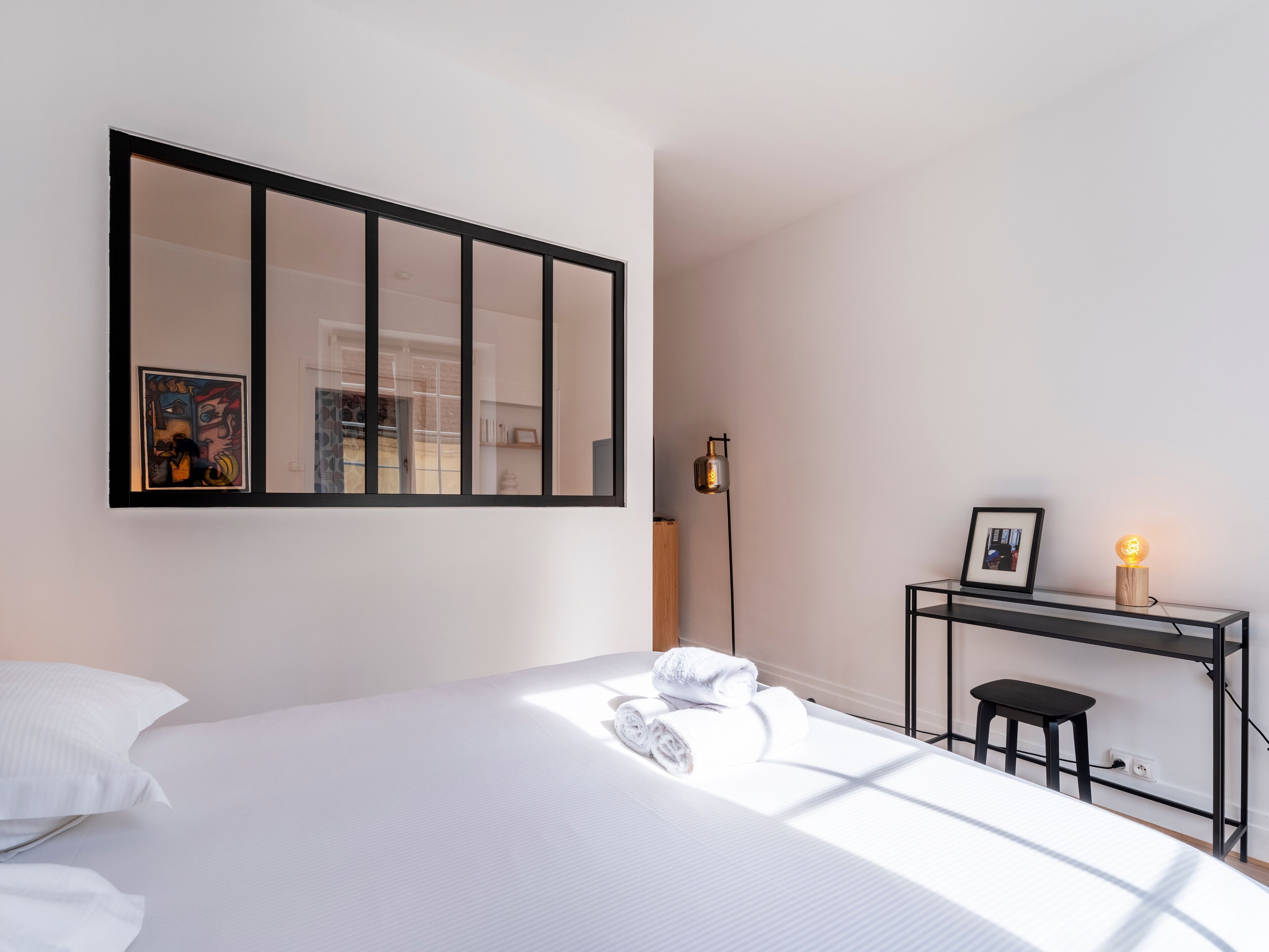 A hotel room with a large bed, a desk with a chair, a standing lamp, and a framed picture on the desk.