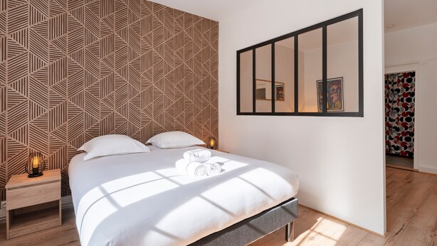 A modern bedroom with a geometric patterned wall, a large bed with white bedding, a bedside table with a lamp, and a mirror on the wall. - L'Alerion Nancéien - Charme au coeur de Nancy (Nancy)