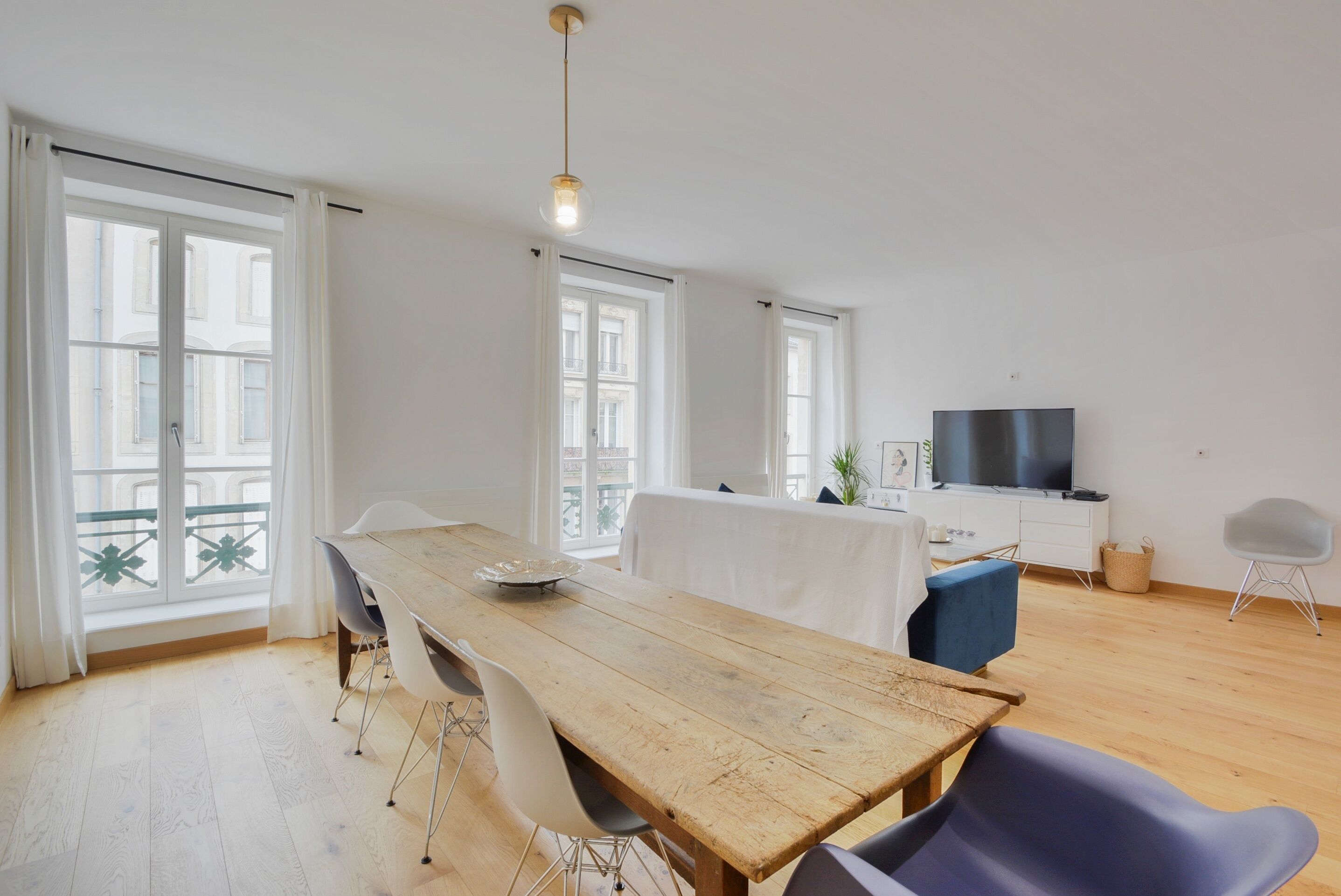 A modern living room with a wooden dining table, white chairs, a TV, and large windows with white curtains.