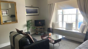 A living room with a sofa, a glass coffee table, a television, a large window with curtains, and a mirror on the wall. - Delightful Howth Village apartment in Dublin (Dublin)