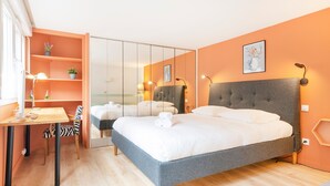 A modern hotel room with a large bed, a desk with a chair, a wardrobe, and a small table with a lamp.