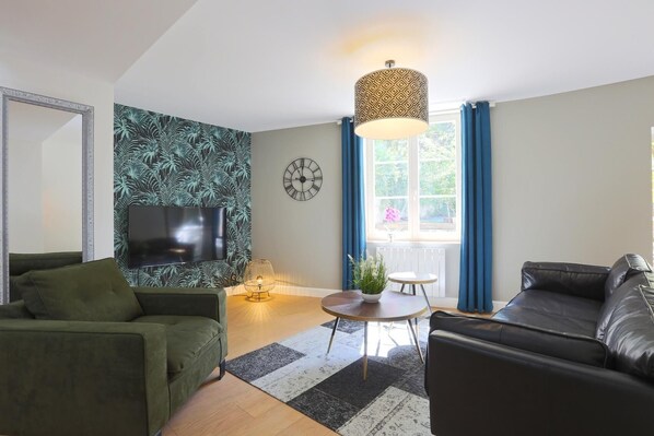 A modern living room with a flat-screen TV, green and black sofas, a round coffee table, and a patterned wall. - Relais Ducal - Côté Jardin (Nancy)