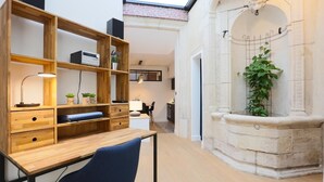 Un espace de bureau moderne avec un bureau en bois, des étagères et une chaise bleue. On aperçoit une fontaine en pierre et une plante à l’arrière-plan.