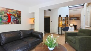 Un salon moderne avec un canapé en cuir noir, un canapé vert et une table basse agrémentée d’un arrangement floral. Un tableau abstrait aux couleurs vives orne le mur, et on aperçoit un coin cuisine à l’arrière-plan.